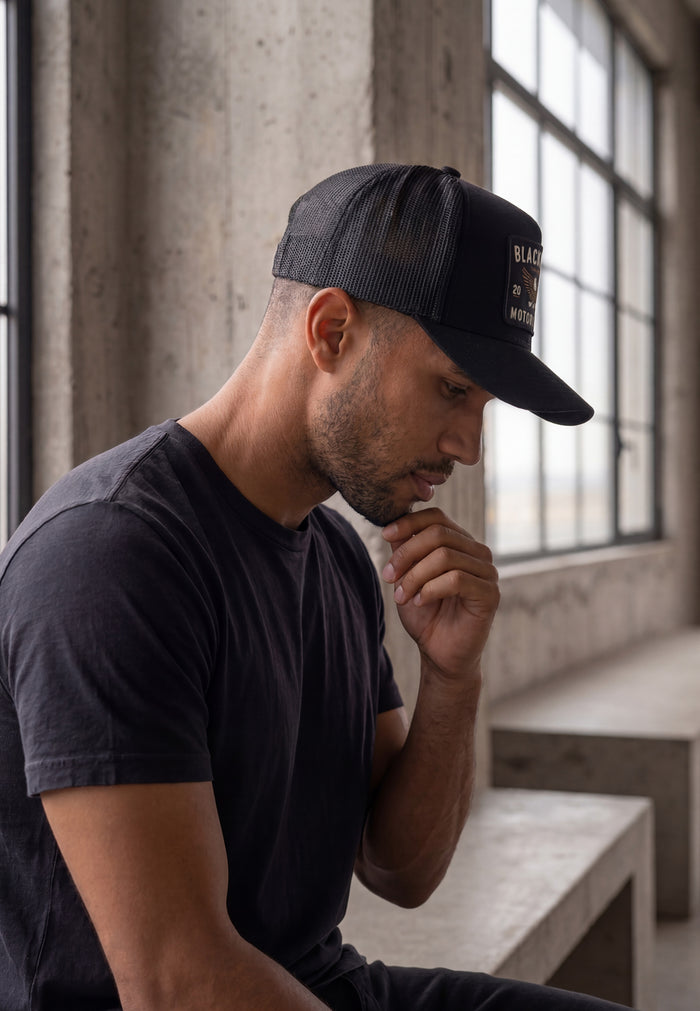 Man wearing a black cap and t-shirt sitting indoors with large windows in the background
