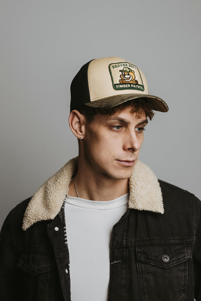 Man wearing a beige and black cap with a logo, black jacket with shearling collar, and white shirt against a gray background.