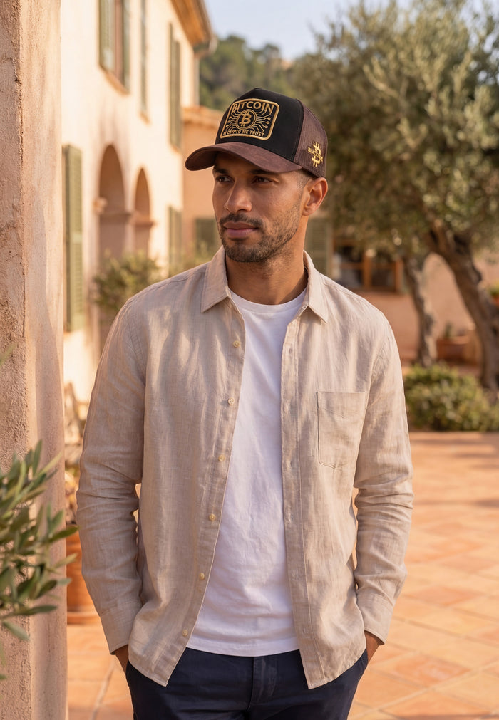Man wearing a bitcoin cap with a logo, beige shirt, and white t-shirt outdoors.