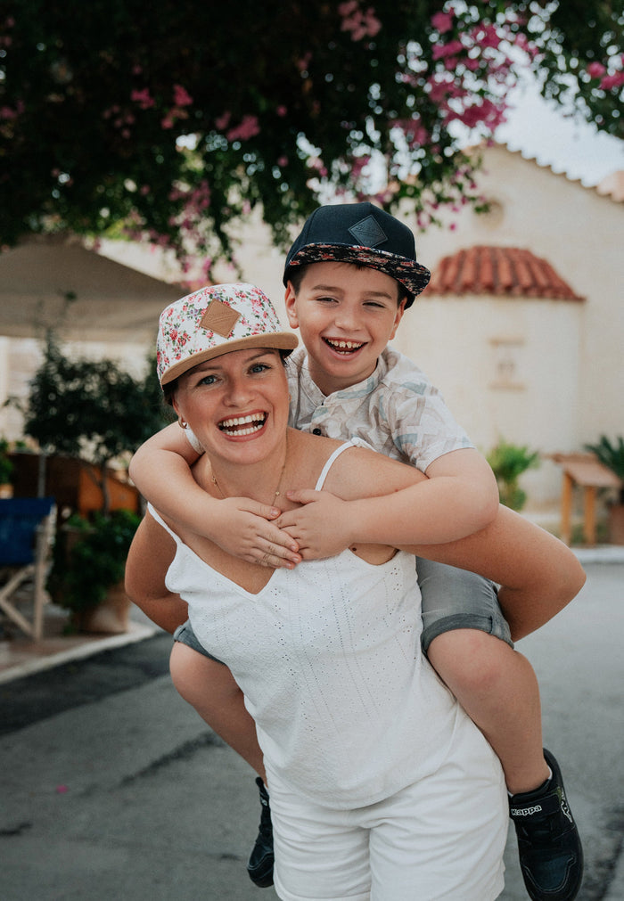 Woman in a white dress giving a piggyback ride to a young boy outdoors with flowers and a building in the background.
