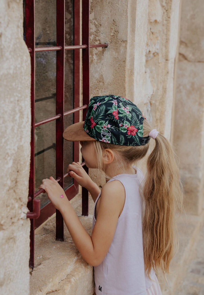 Young girl wearing a floral cap and white sleeveless top, standing in front of a stone wall with a red metal gate.