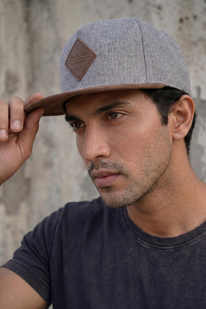 Man wearing a gray cap with a brown leather patch against a textured wall.