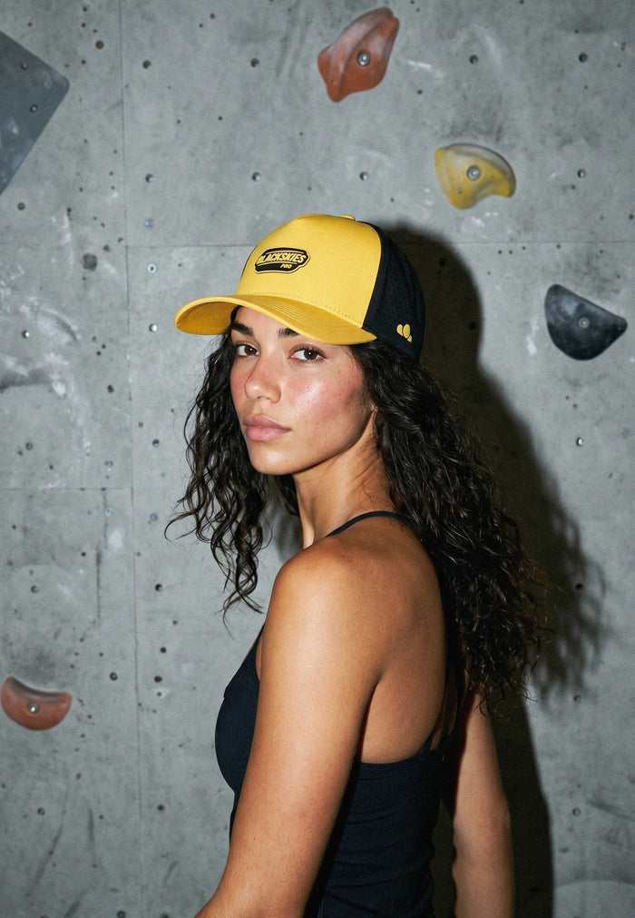 Woman wearing a yellow cap with a logo against a climbing wall background