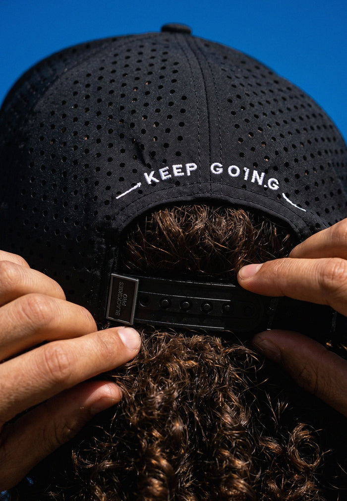 Black cap with 'KEEP GOING' text held by a person against a blue background