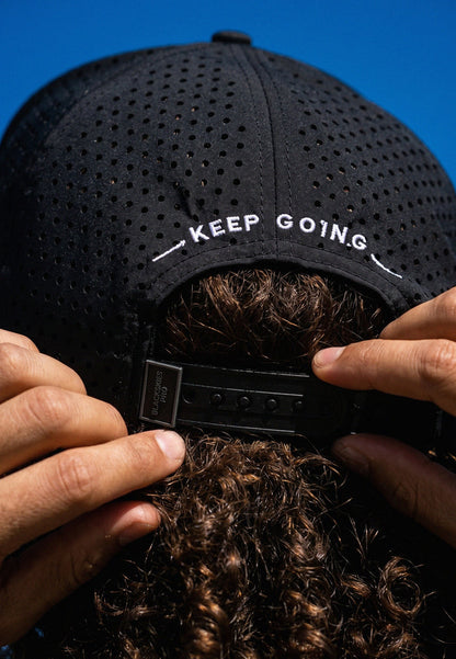 Black cap with 'KEEP GOING' text held by a person against a blue background