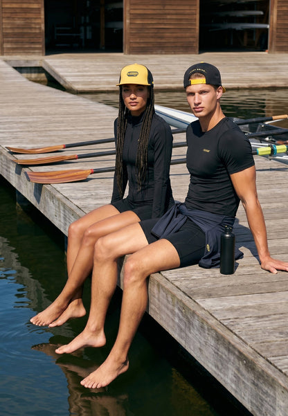 Two people sitting on a dock by a body of water, wearing black athletic wear with yellow accents.
