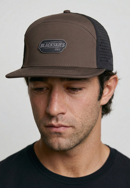 Man wearing a brown cap with a visible brand logo on a plain background