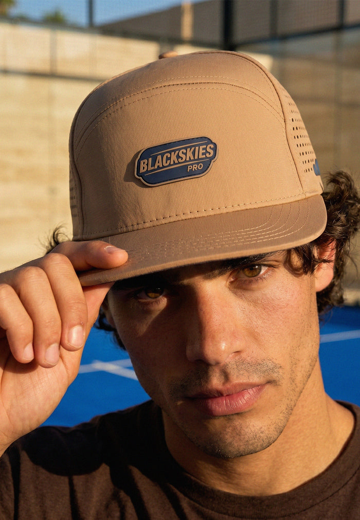 Man wearing a tan cap with 'Blackskies Pro' logo against a blue and gray background