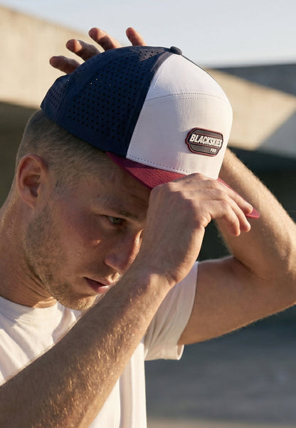 Man wearing a navy, white, and red cap with 'BlackStrap' branding on a blurred beach background.