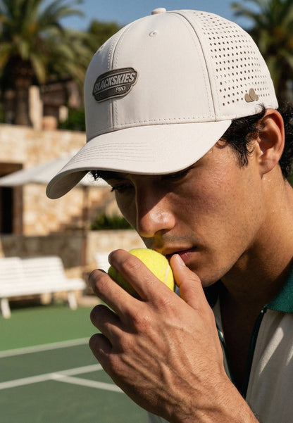 Man holding a tennis ball on a tennis court wearing a cap with a brand logo.