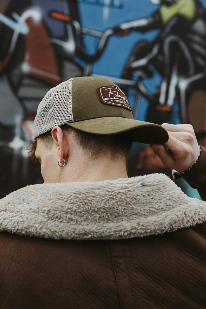 Person wearing a brown cap with a logo, adjusting it against a graffiti-covered wall.
