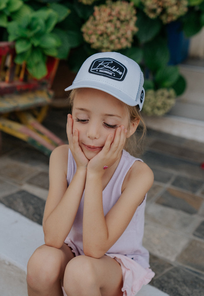 Young girl wearing a white cap with a logo, sitting on a stone floor with plants in the background.