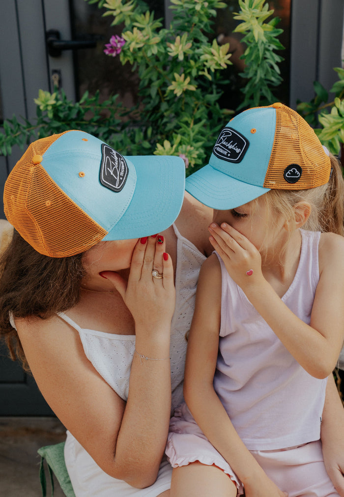 Woman and child wearing matching blue and orange caps outdoors with greenery in the background