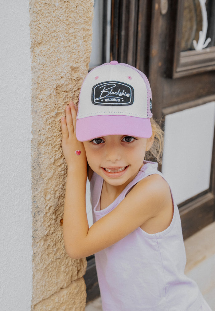 Young girl wearing a pink and white cap with a logo, standing against a textured wall.