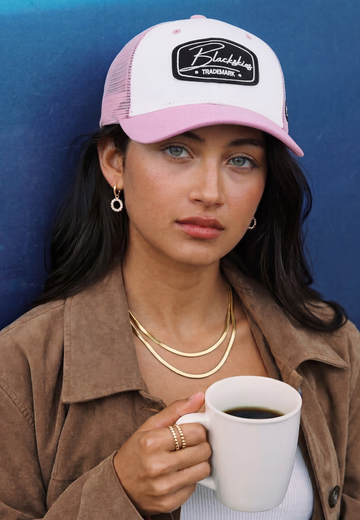 Woman wearing a pink and white blackskies trucker cap with a logo, holding a coffee cup against a blue background