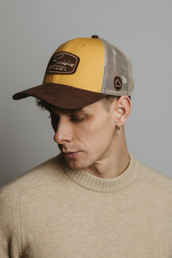 Man wearing a mustard yellow and brown cap with a visible brand logo on a gray background