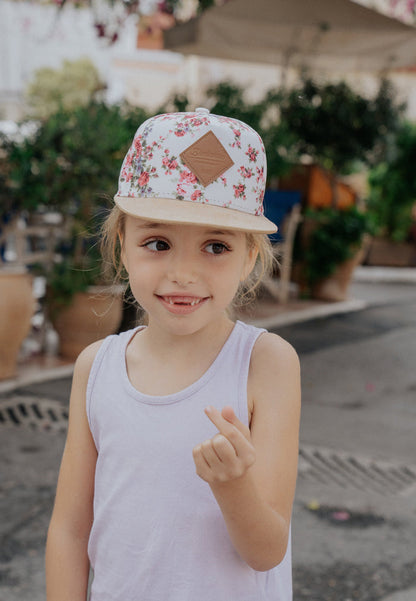 Young girl wearing a floral cap and white tank top, standing outdoors with plants and an umbrella in the background.