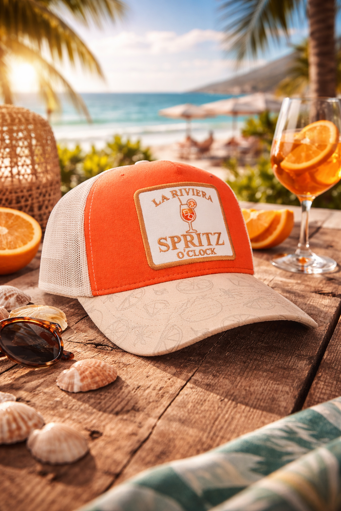 Orange and beige cap with 'La Riviera Spritz O'Clock' text on a wooden table by the beach.